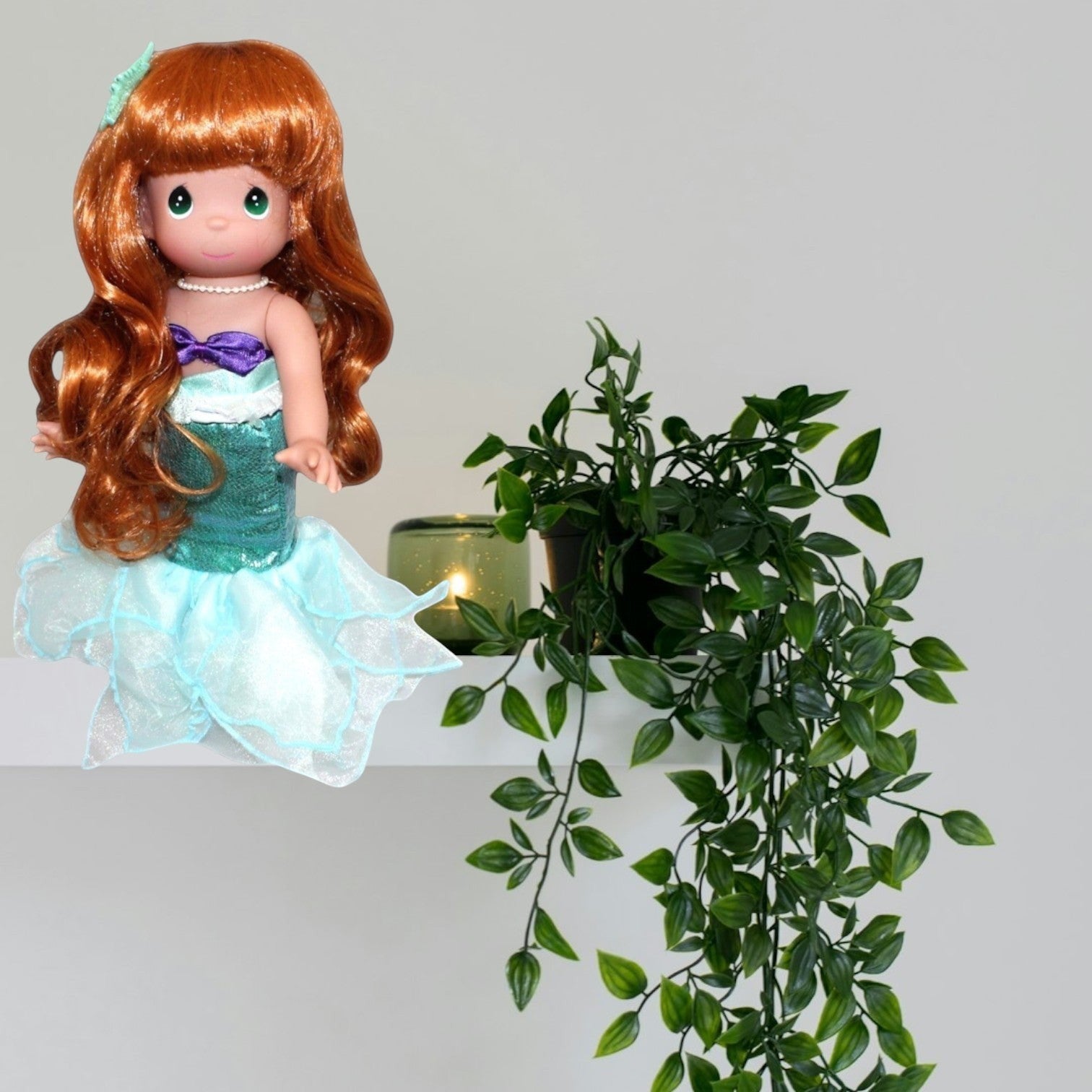 The Doll Maker Ariel mermaid doll with long red hair and a teal shimmering tail sitting on a white shelf with a plant