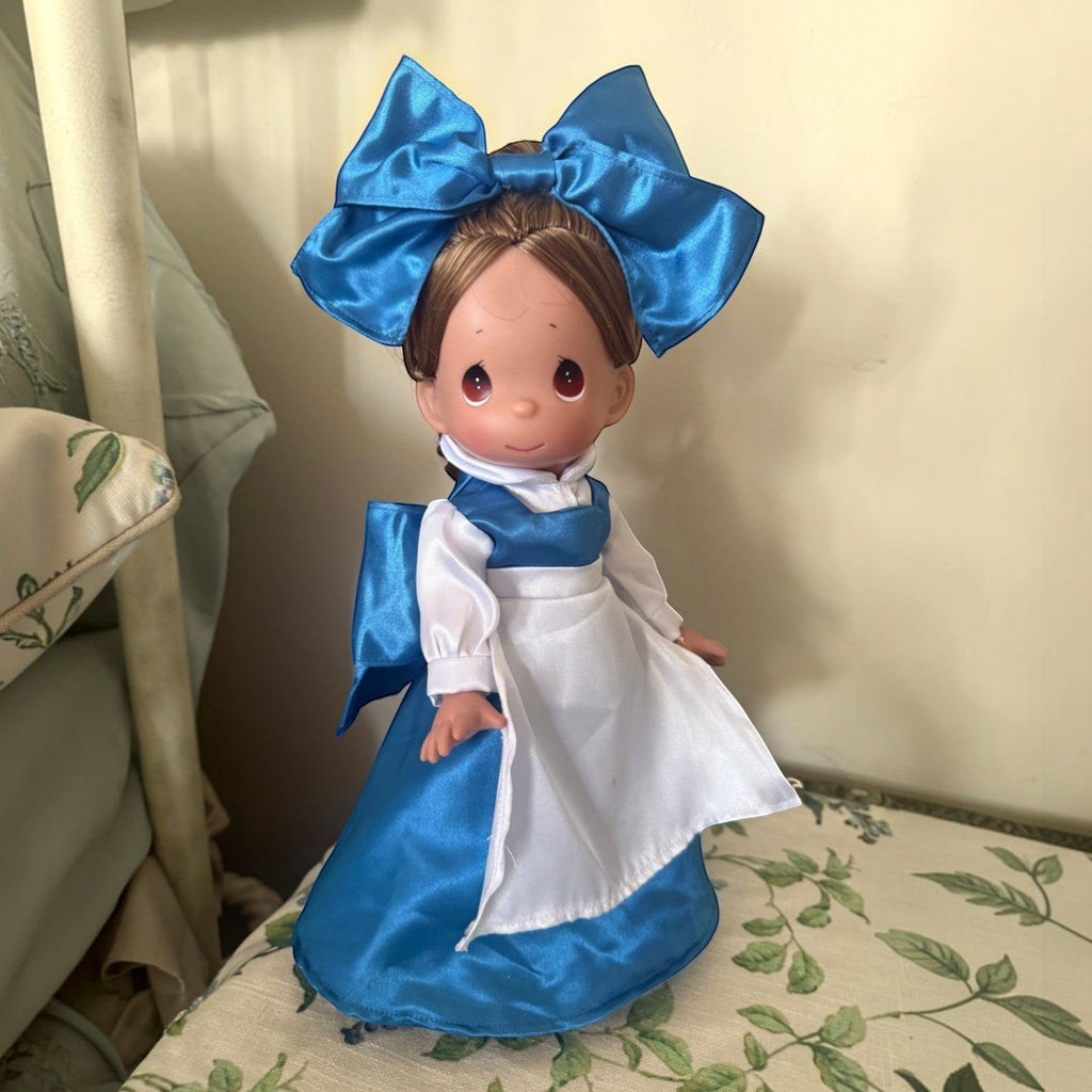 The Doll Maker 12-inch Belle doll wearing a blue dress with a white apron and a large matching blue hair bow