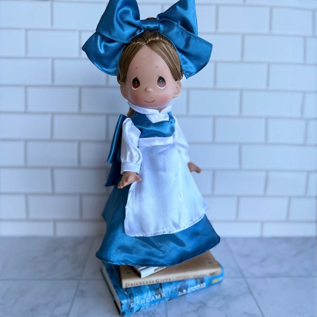The Doll Maker 12-inch Belle doll wearing a blue dress with a white apron and a large matching blue hair bow.