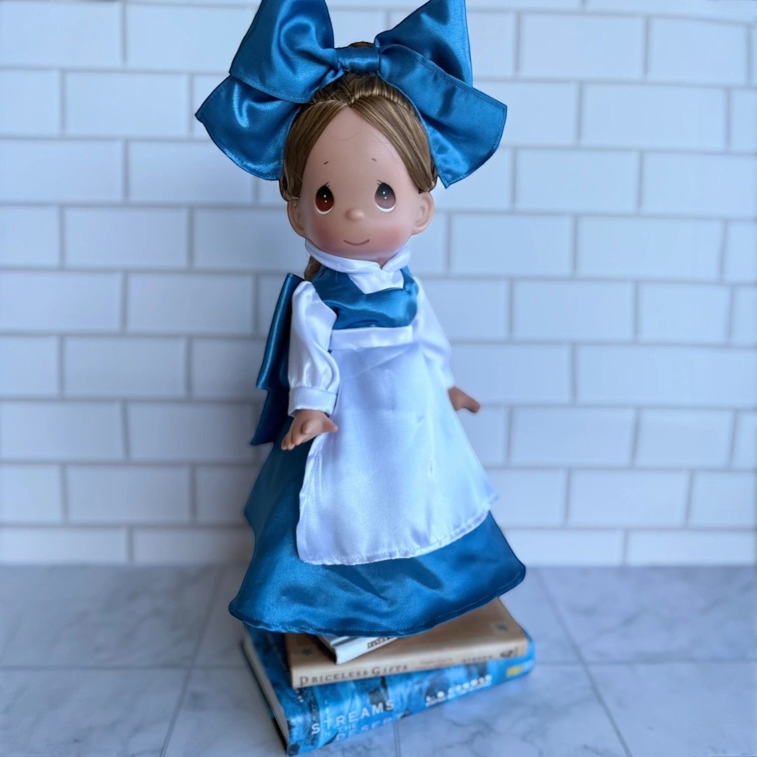 The Doll Maker 12-inch Belle doll wearing a blue dress with a white apron and a large matching blue hair bow.