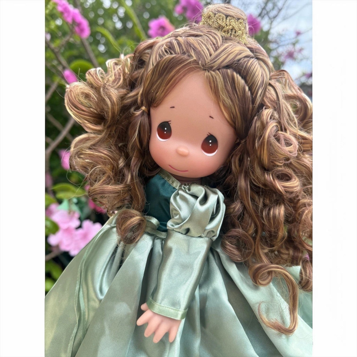 The Doll Maker 12-inch Beautiful Belle doll with curly brown hair and sage green satin gown in a garden setting.