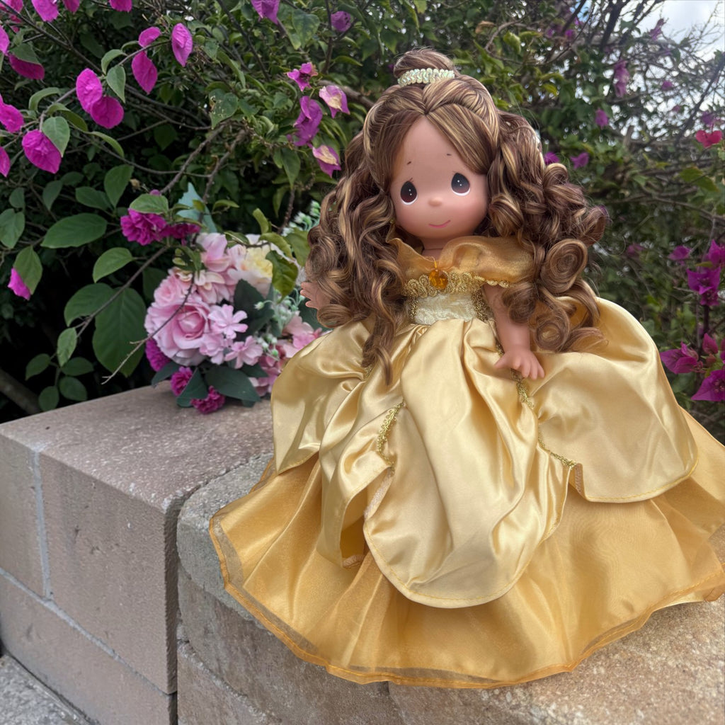 The Doll Maker 12-inch Belle doll wearing an elegant gold gown with a matching tiara in a garden setting.