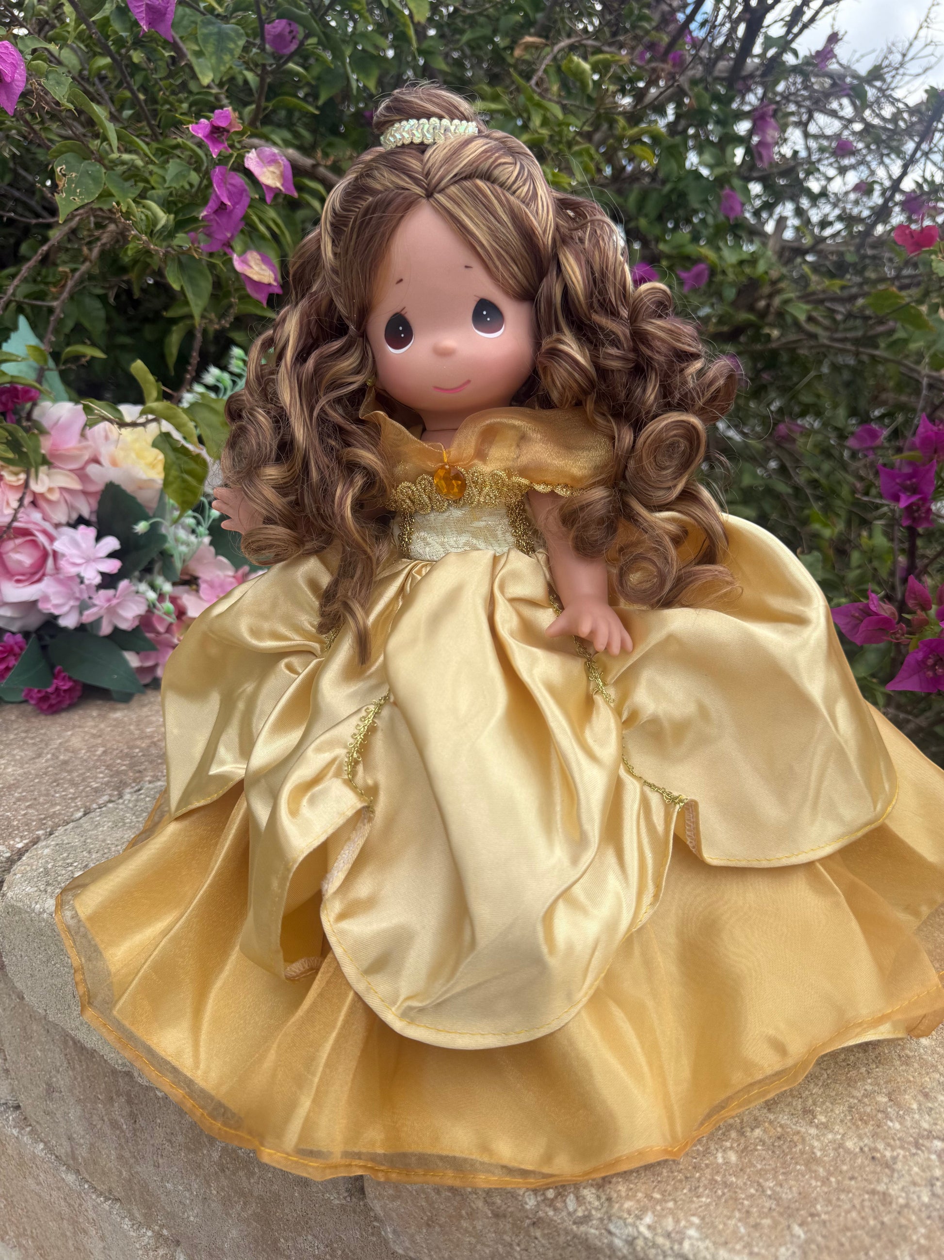 The Doll Maker 12-inch Belle doll wearing an elegant gold gown with a matching tiara in a garden setting.