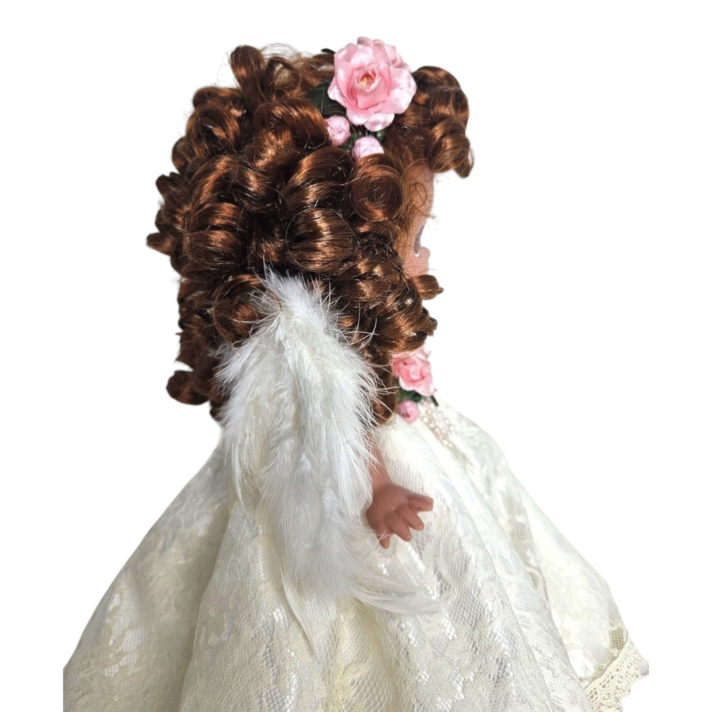 The Doll Maker 12-inch brunette angel doll wearing a white lace dress with feathered wings and pink floral hair decor.