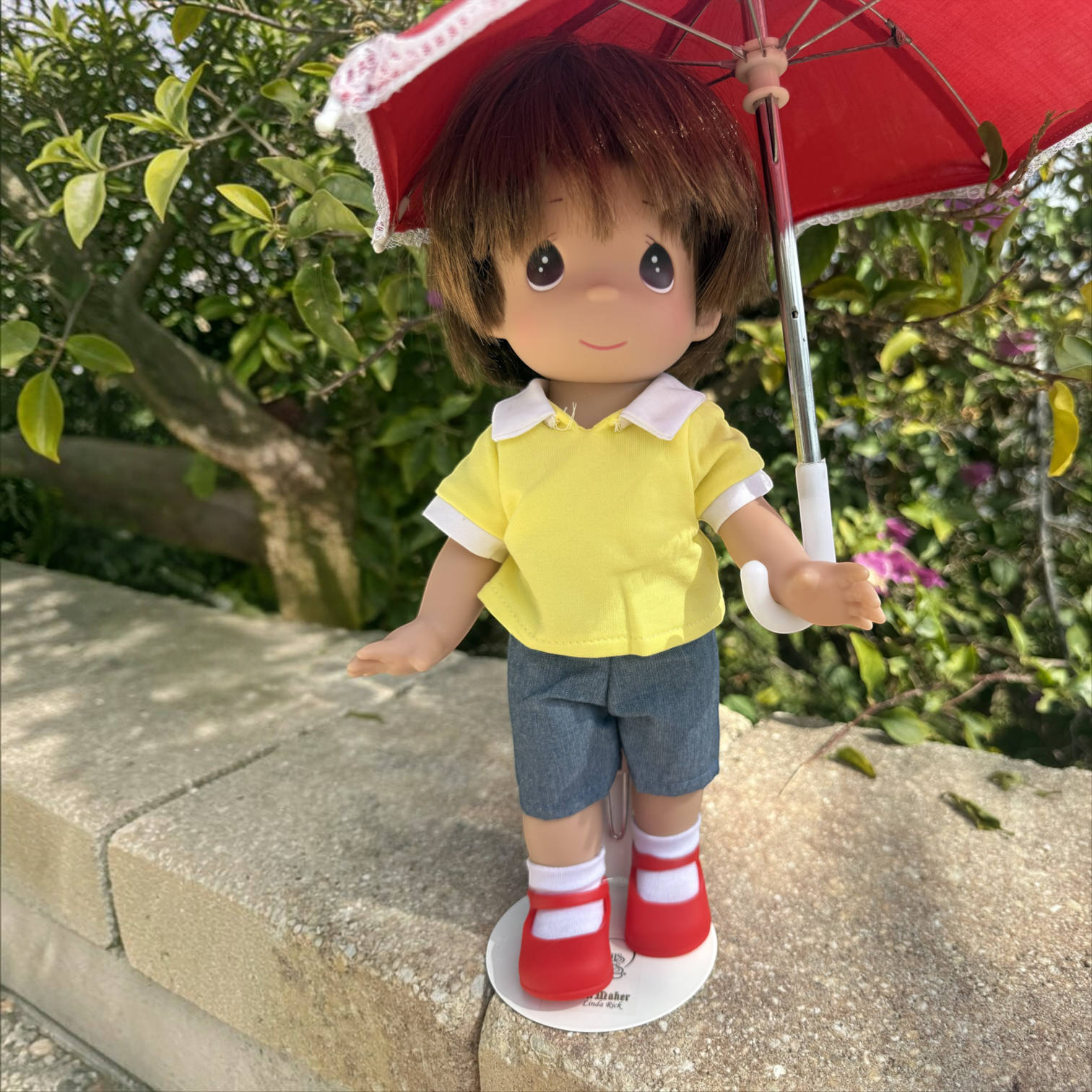 The Doll Maker 12-inch Christopher Robin doll wearing a yellow shirt and blue shorts, holding a red umbrella outdoors.
