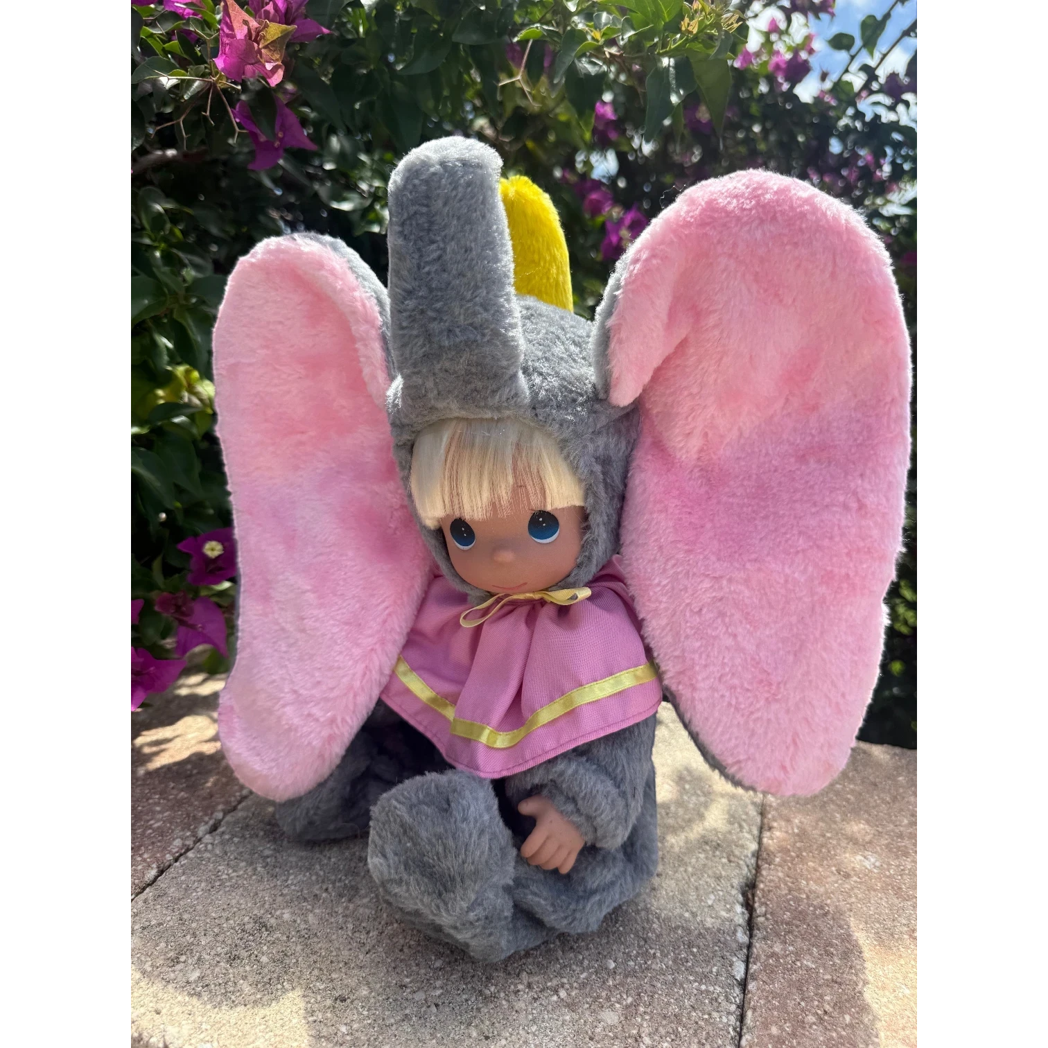 The Doll Maker Dumbo sitting doll with large pink plush ears, grey fuzzy elephant costume, and a yellow hat.