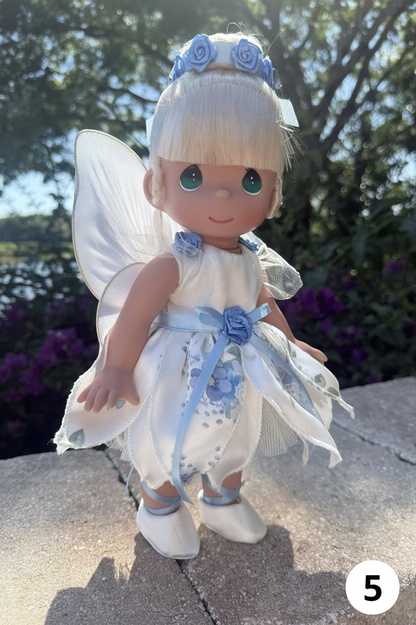 The Doll Maker 12-inch princess fairy doll with blonde hair, blue floral dress, and sheer wings in a garden setting.