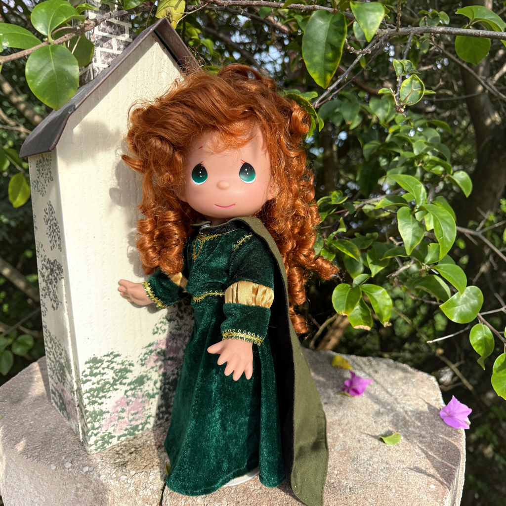 The Doll Maker 12-inch Merida doll from Brave with curly red hair wearing a green velvet dress outdoors.