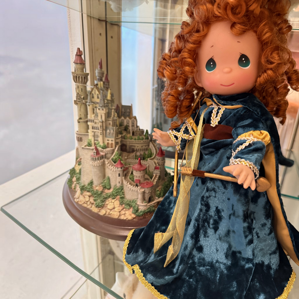 The Doll Maker Merida Brave warrior doll with curly red hair in a blue velvet dress standing beside a castle model.