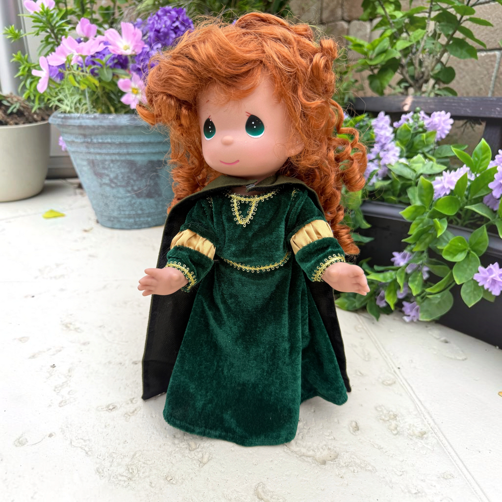 The Doll Maker 12-inch Merida doll with curly red hair wearing a green velvet dress and cape in a garden setting.