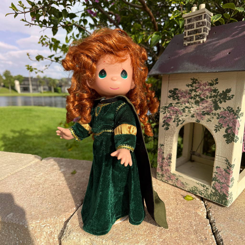The Doll Maker 12-inch Merida doll with curly red hair wearing a forest green velvet dress and cape outdoors.