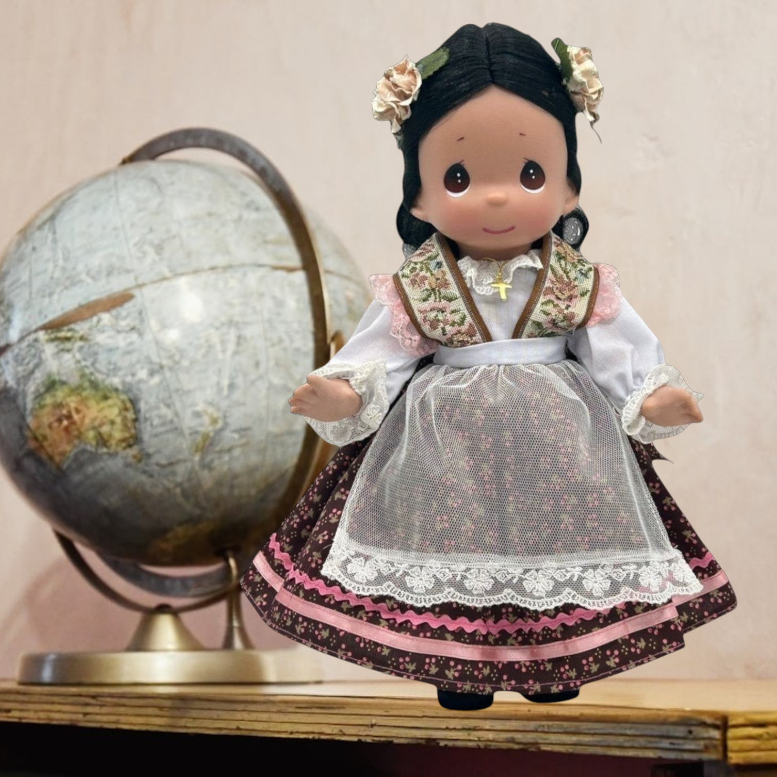 The Doll Maker 12-inch Mexico doll wearing a traditional floral dress and lace apron with a globe in the background.