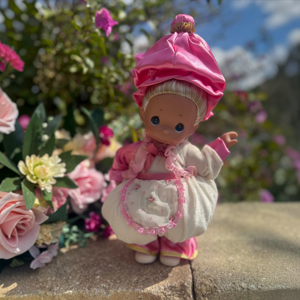 The Doll Maker Mrs. Potts 12-inch collectible doll wearing a pink satin hat and floral apron in a garden setting.