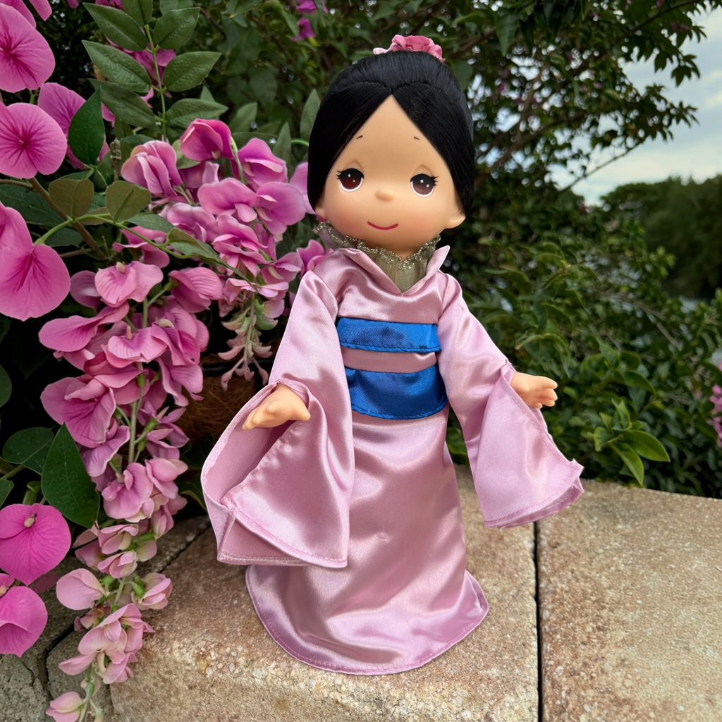 The Doll Maker Mulan 12-inch vinyl doll wearing a pink satin kimono with a blue obi belt, posed by pink flowers.