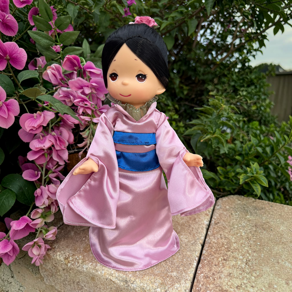 The Doll Maker 12-inch Mulan vinyl doll wearing a pink satin robe with a blue sash, standing near pink flowers.