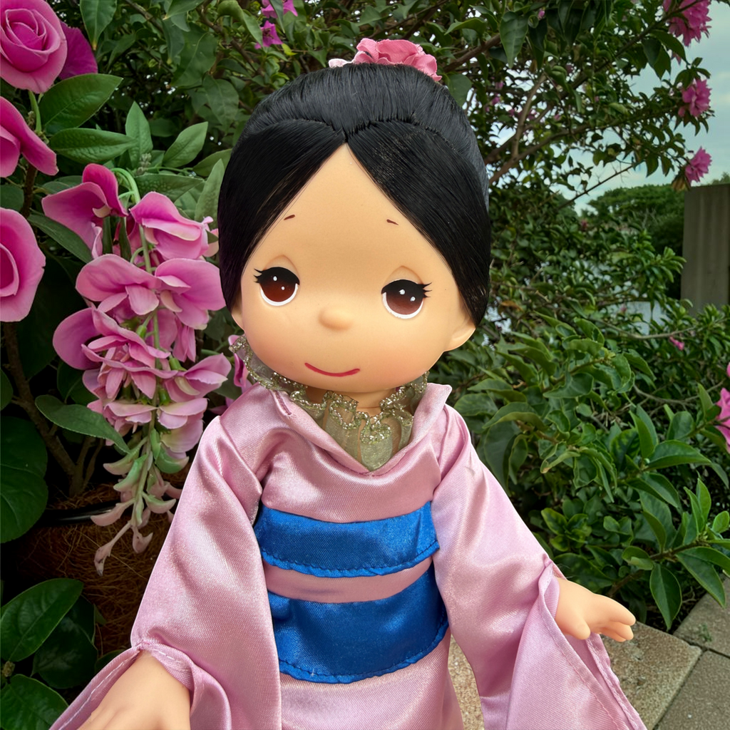 The Doll Maker Mulan 12-inch vinyl doll wearing pink robes with a blue sash, posed outdoors near pink flowers.