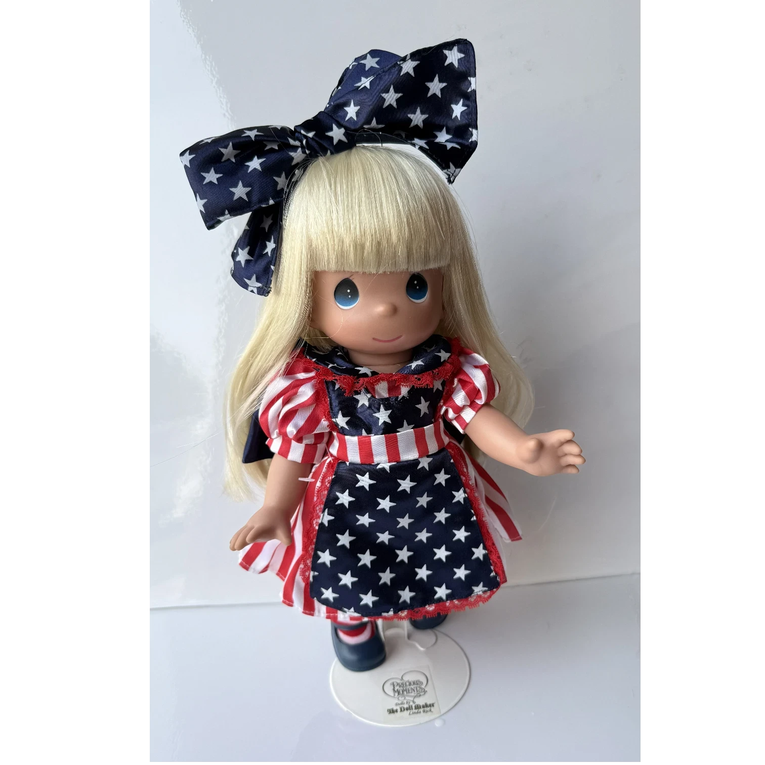 The Doll Maker Patriotic Alice 12-inch collectible doll with blonde hair, star-print dress, and matching hair bow.