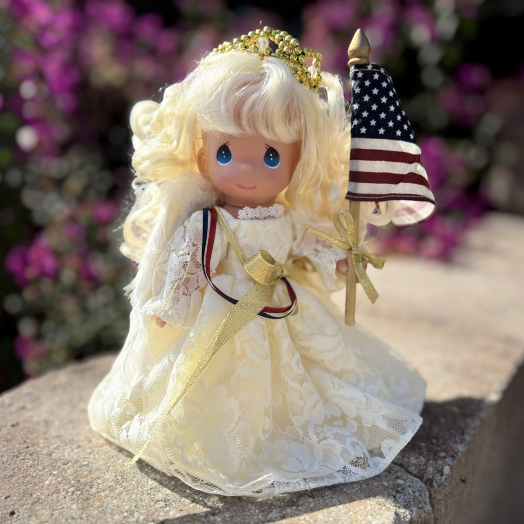 The Doll Maker 9-inch Sweet Liberty angel doll wearing a white lace gown with a gold crown and holding a US flag.