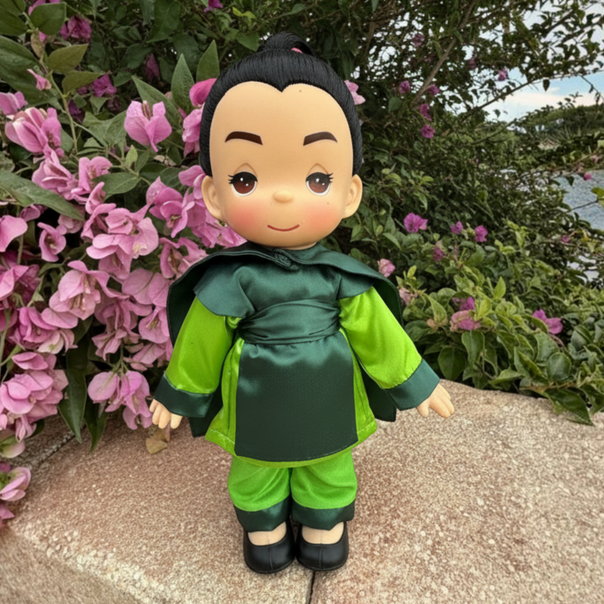 The Doll Maker Ping 12-inch vinyl doll wearing green warrior armor, standing outdoors against pink bougainvillea.
