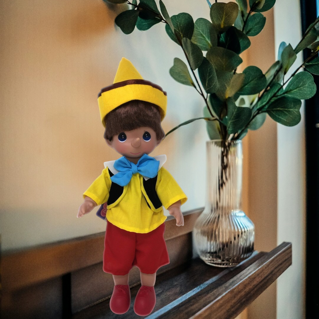 The Doll Maker 12-inch Pinocchio doll wearing a yellow hat, yellow shirt, blue bow tie, and red shorts on a shelf.