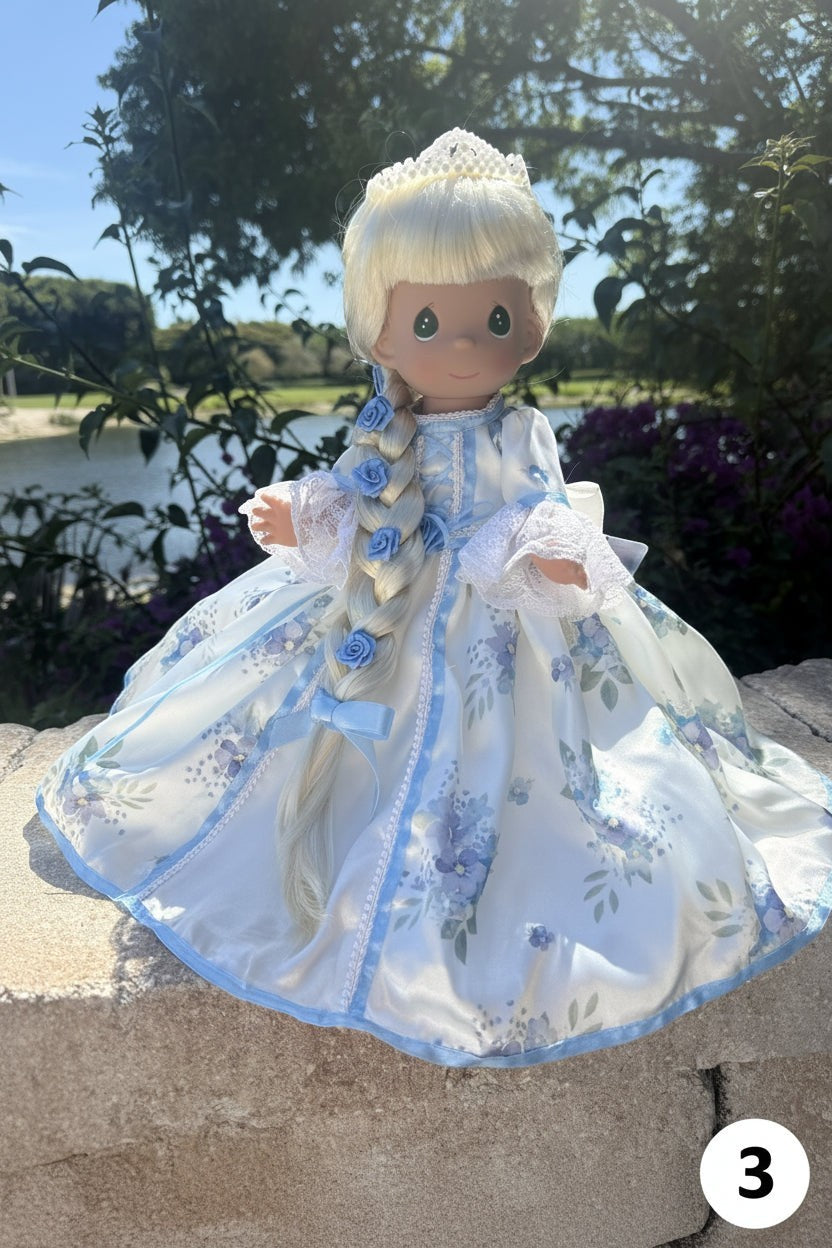 The Doll Maker 12-inch princess doll with blonde braided hair, blue floral satin gown, and tiara in a garden setting.