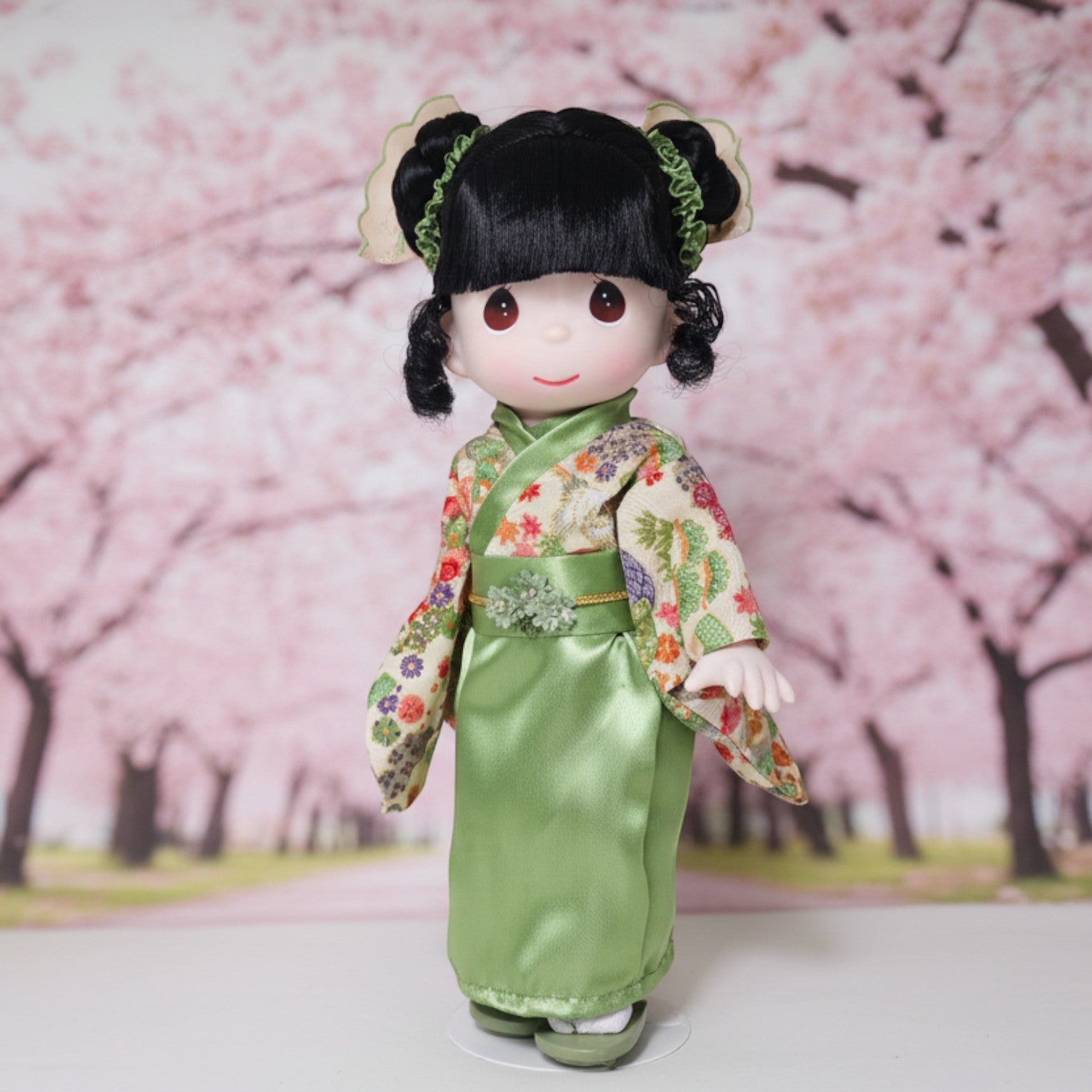 The Doll Maker Rui doll wearing a green floral kimono for Japanese Girls' Day, standing before a cherry blossom backdrop