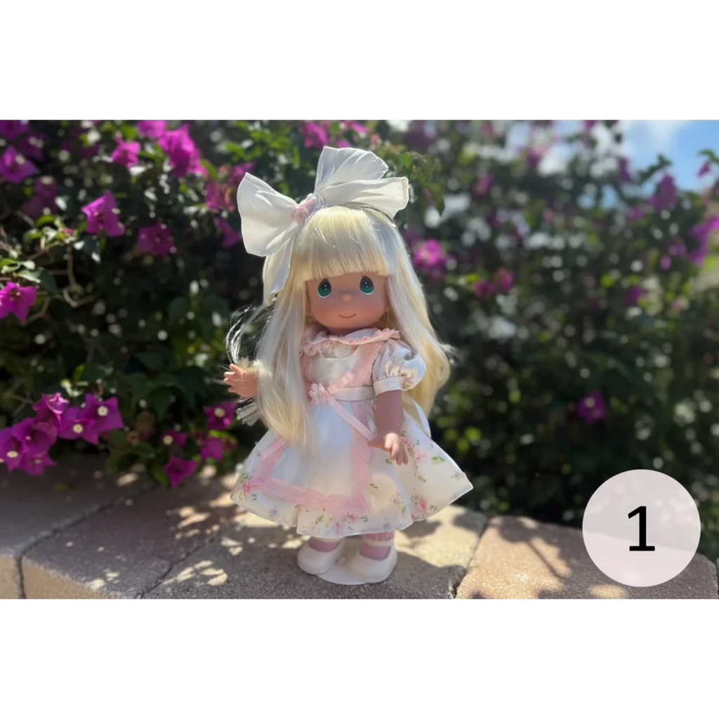 The Doll Maker 12-inch Spring Bouquets doll with blonde hair, wearing a floral dress and large white hair bow.
