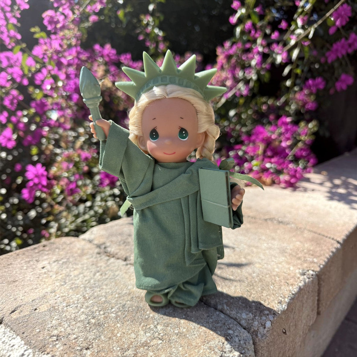 The Doll Maker 7-inch Statue of Liberty vinyl doll with blonde hair and green robe standing on a stone wall.