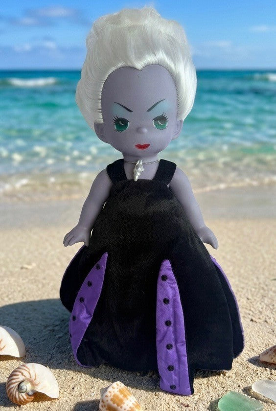The Doll Maker 12-inch Ursula vinyl doll with white hair and black and purple dress standing on a sandy beach.