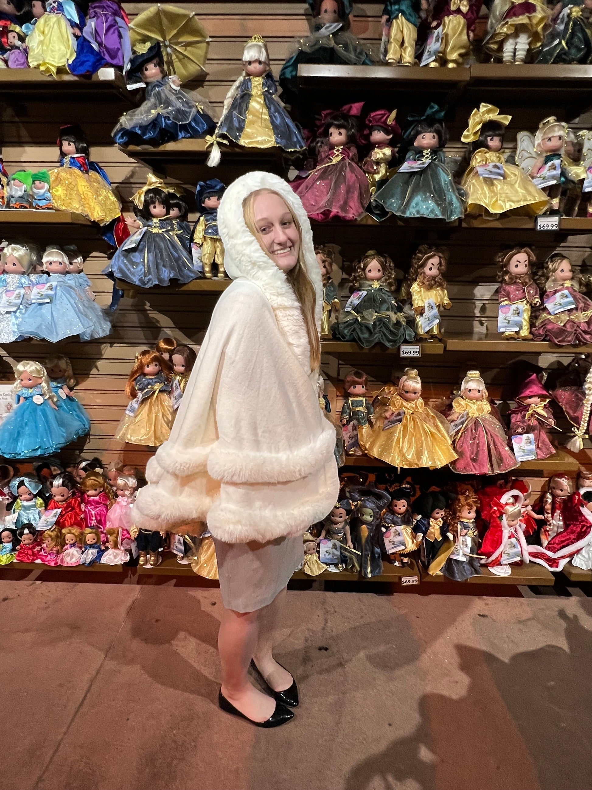 A woman wearing a white hooded fur-trimmed cape dresses like Linda Rick, standing in a store filled with collectible dolls Linda created