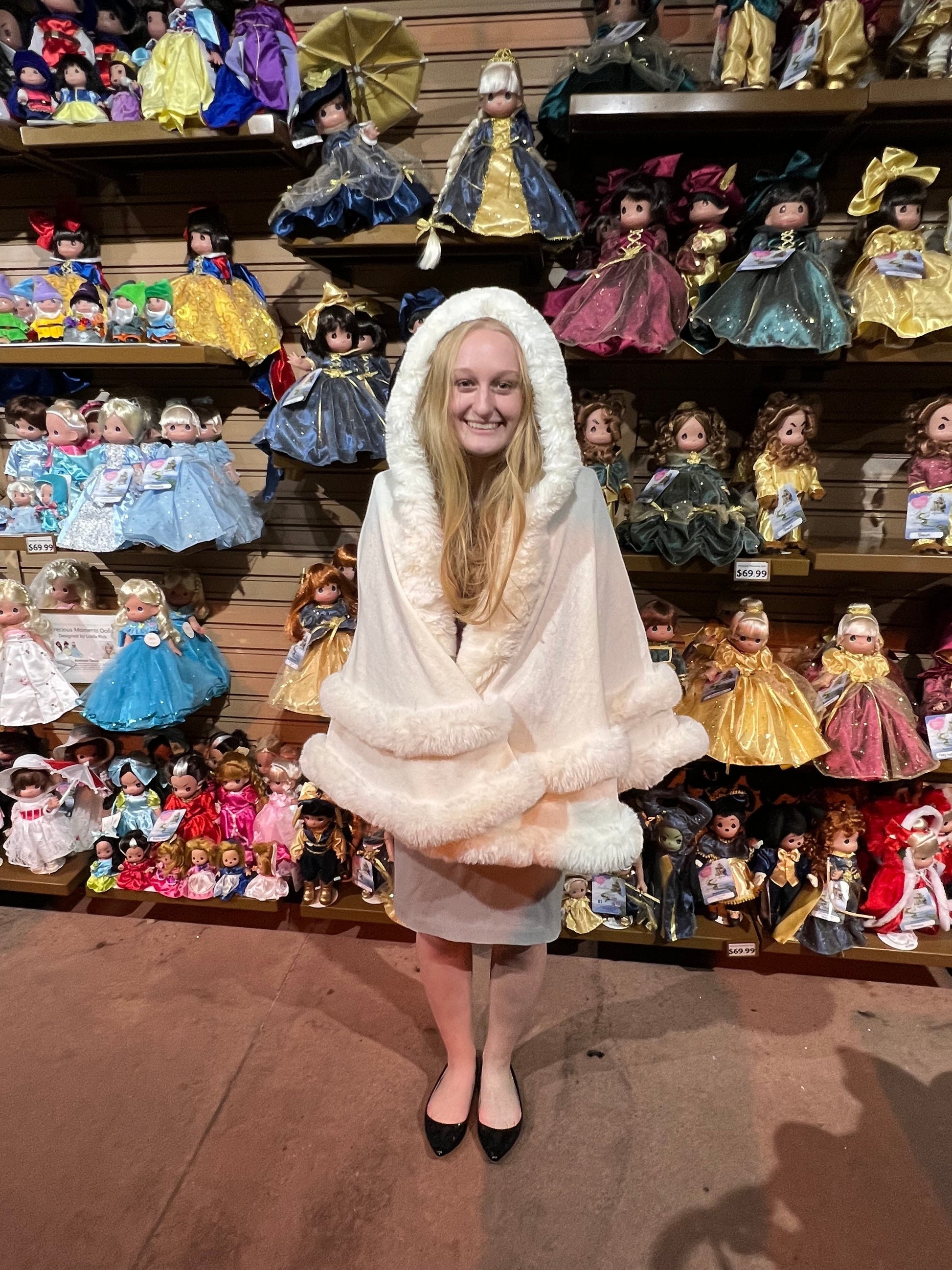 White hooded winter cape with plush fur trim worn by a smiling woman in a doll shop.