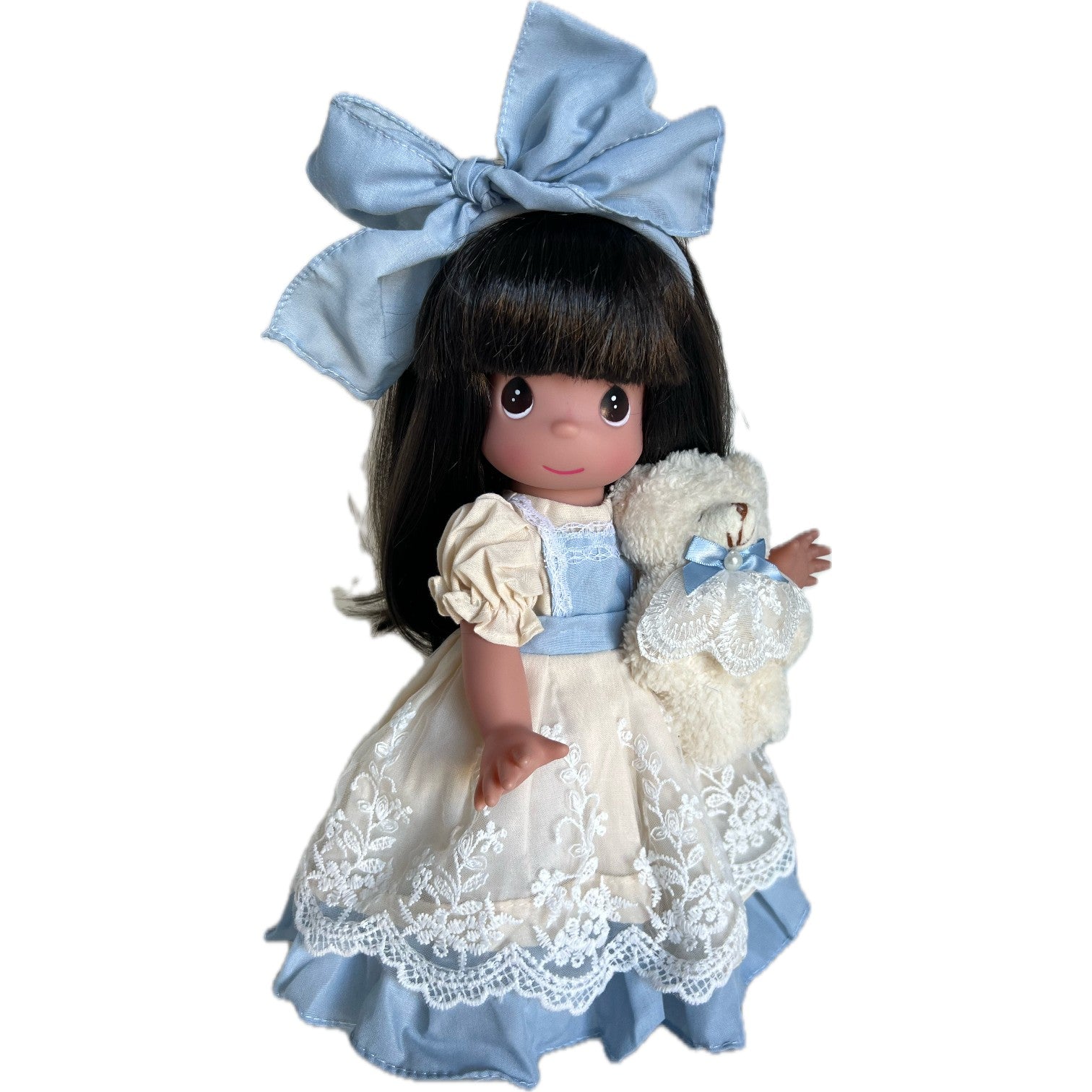 The Doll Maker 12" vinyl doll with brunette hair, blue bow, and a cream dress with blue trim and lace details, holding a teddy bear.