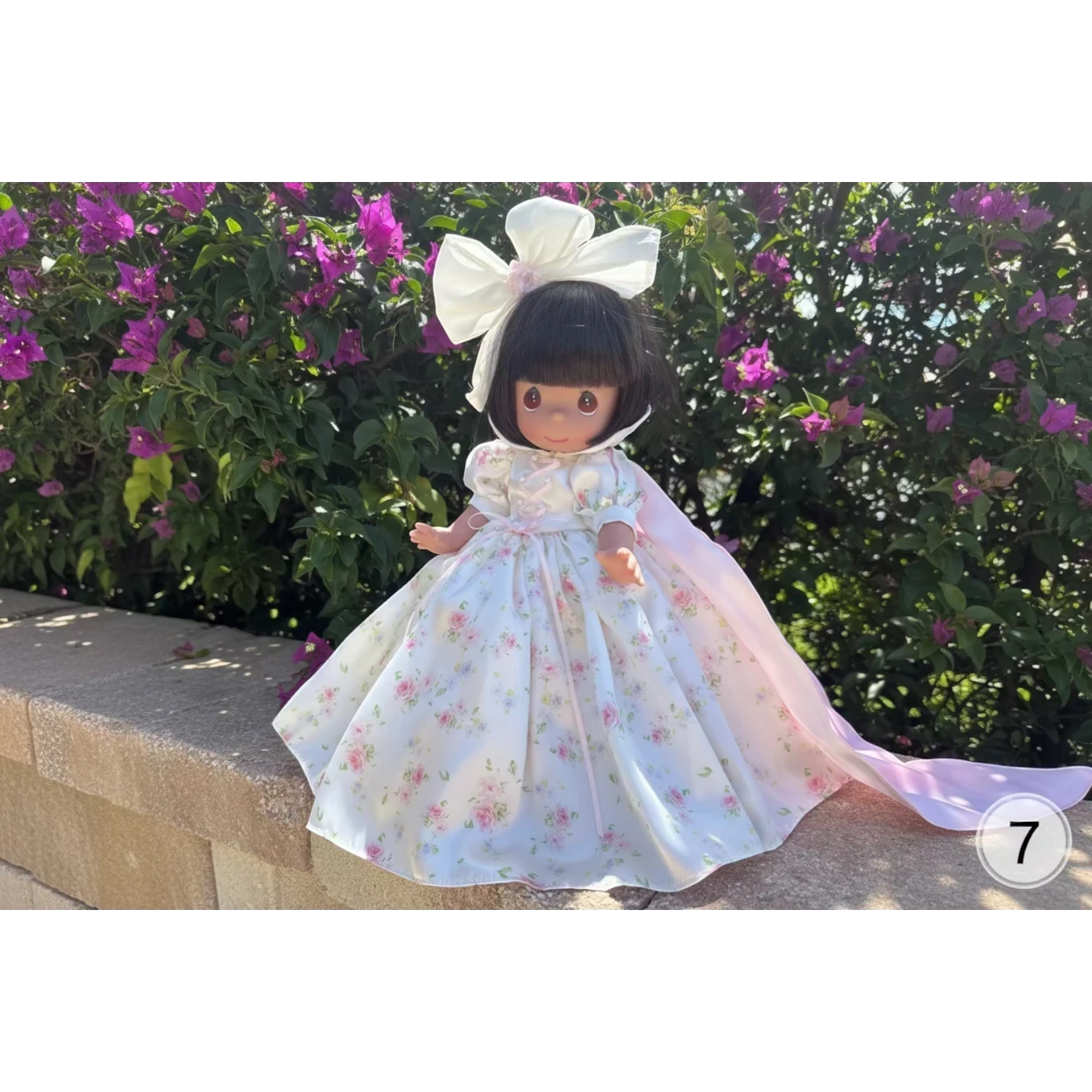 The Doll Maker's 12-inch Spring Bouquets doll wears a white floral dress with a pink sash and a matching pink cape. Features a large white bow on her head.