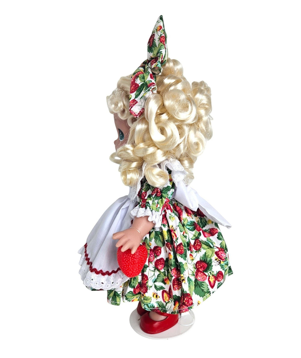 The Doll Maker's You’re Berry Sweet 12” doll with blonde curly hair, wearing a strawberry print dress and holding a strawberry.